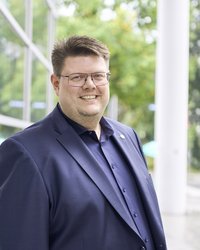 Foto von Jörg Fritzsche im blauen Jacket mit Anstecknadel mit dem Sonnenblumenlogo der Grünen vor dem Hagener Rathaus