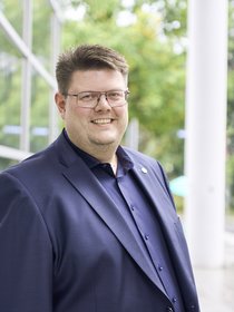 Foto von Jörg Fritzsche im blauen Jacket mit Anstecknadel mit dem Sonnenblumenlogo der Grünen vor dem Hagener Rathaus