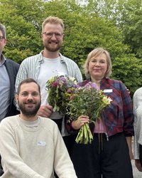 Gruppenfoto mit fünf Personen im Freien, die in die Kamera lächeln. Im Hintergrund sieht man grüne Laubbäume. Die zweite Person von links sitzt in einem Rollstuhl, alle anderen stehen. Die beiden Personen in der Mitte haben Blumensträuße in der Hand.