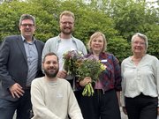 Gruppenfoto mit fünf Personen im Freien, die in die Kamera lächeln. Im Hintergrund sieht man grüne Laubbäume. Die zweite Person von links sitzt in einem Rollstuhl, alle anderen stehen. Die beiden Personen in der Mitte haben Blumensträuße in der Hand.