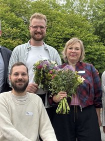 Gruppenfoto mit fünf Personen im Freien, die in die Kamera lächeln. Im Hintergrund sieht man grüne Laubbäume. Die zweite Person von links sitzt in einem Rollstuhl, alle anderen stehen. Die beiden Personen in der Mitte haben Blumensträuße in der Hand.